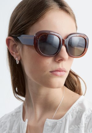 Młoda kobieta nosząca duże brązowe przezroczyste okulary przeciwsłoneczne, srebrne kolczyki-koła, delikatny naszyjnik i białą haftowaną bluzkę.