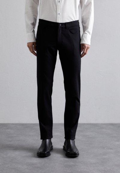 Pantalon noir en tissu stretch lisse. Présente un design ajusté, des poches avant et une fermeture à bouton. Porté avec une chemise légère à boutons.