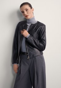 Kobieta ubrana w szary golf, czarną skórzaną kurtkę i szare szerokie spodnie w prążki, stojąca na tle jasnej ściany.