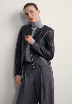 Kobieta ubrana w szary golf, czarną skórzaną kurtkę i szare szerokie spodnie w prążki, stojąca na tle jasnej ściany.