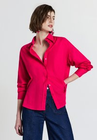 Donna che indossa una camicia button-up rosa vivace parzialmente infilata in un paio di jeans blu scuro, in piedi con una mano sul fianco contro uno sfondo bianco.