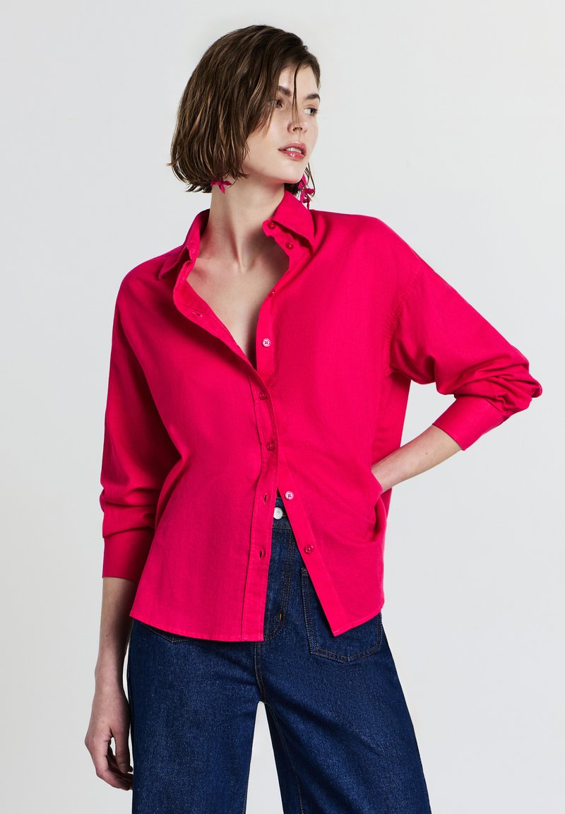 Donna che indossa una camicia button-up rosa vivace parzialmente infilata in un paio di jeans blu scuro, in piedi con una mano sul fianco contro uno sfondo bianco.