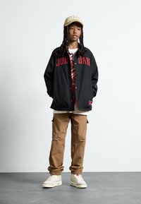 Svart jakke med rød "JORDAN" tekst, beige caps, grafisk t-skjorte, brune bukser og hvite sneakers med grå detaljer. Jevne teksturer gjennom hele antrekket.