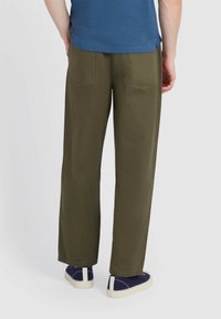 Olijfgroene wijde broek met twee achterzakken, gedragen met een blauw shirt en marineblauwe schoenen met witte zolen.