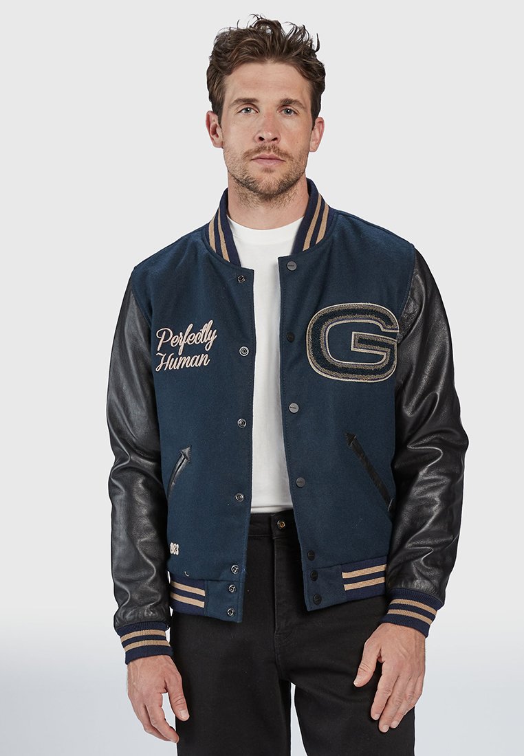 Blauwe en zwarte varsityjack met leren mouwen, geribbelde kraag, geborduurde tekst "Perfectly Human" en gouden accenten. Beschikt over een drukknopen sluiting aan de voorkant.