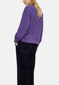 Fioletowy sweter z prążkowanym dołem i mankietami, zestawiony z ciemnymi dżinsami z kontrastowym przeszyciem. Gładka tekstura, luźny krój.