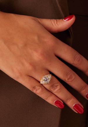 Gouden ring in marquise-vorm met een centrale heldere steen omringd door kleinere stenen, gedragen op een hand met rode nagellak.