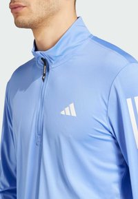 Light blue training jacket with a half-zip collar, featuring a white logo on the chest and striped accents on the shoulders. Smooth fabric texture.