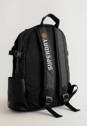 Čierny batoh s polstrovanými popruhmi, nastaviteľnými prackami a prednou zipsovou kapsou. Obsahuje logo "SUPERDRY" v bielej farbe a oranžový akcent.
