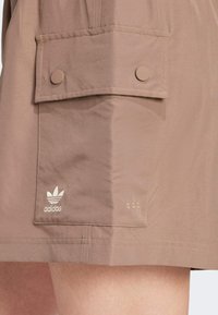 Brown athletic skirt made from smooth material, featuring a side pocket with button closure and the Adidas logo in white embroidery.