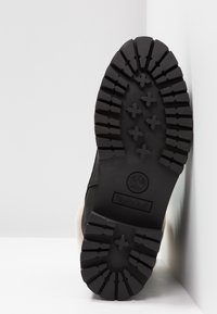 Semelle de botte Timberland noire avec des crampons profonds et un logo, positionnée verticalement contre une surface et un mur blancs.