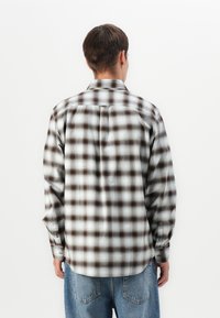 Mand iført en langærmet, knappet, grå og brun plaid skjorte og blå jeans, set bagfra mod en hvid baggrund.