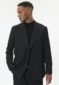 Juodos dvigubos krūtinės švarkas, pagamintas iš tekstūruotos medžiagos, su nupjautomis apykaklėmis ir juodais mygtukais. Nešiojamas ant juodos golfos. Švarios linijos ir pritaikytas siluetas.