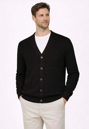 Uomo con cardigan nero abbottonato sopra una camicia bianca e pantaloni beige chiaro, in piedi con le mani nelle tasche, che guarda di lato.