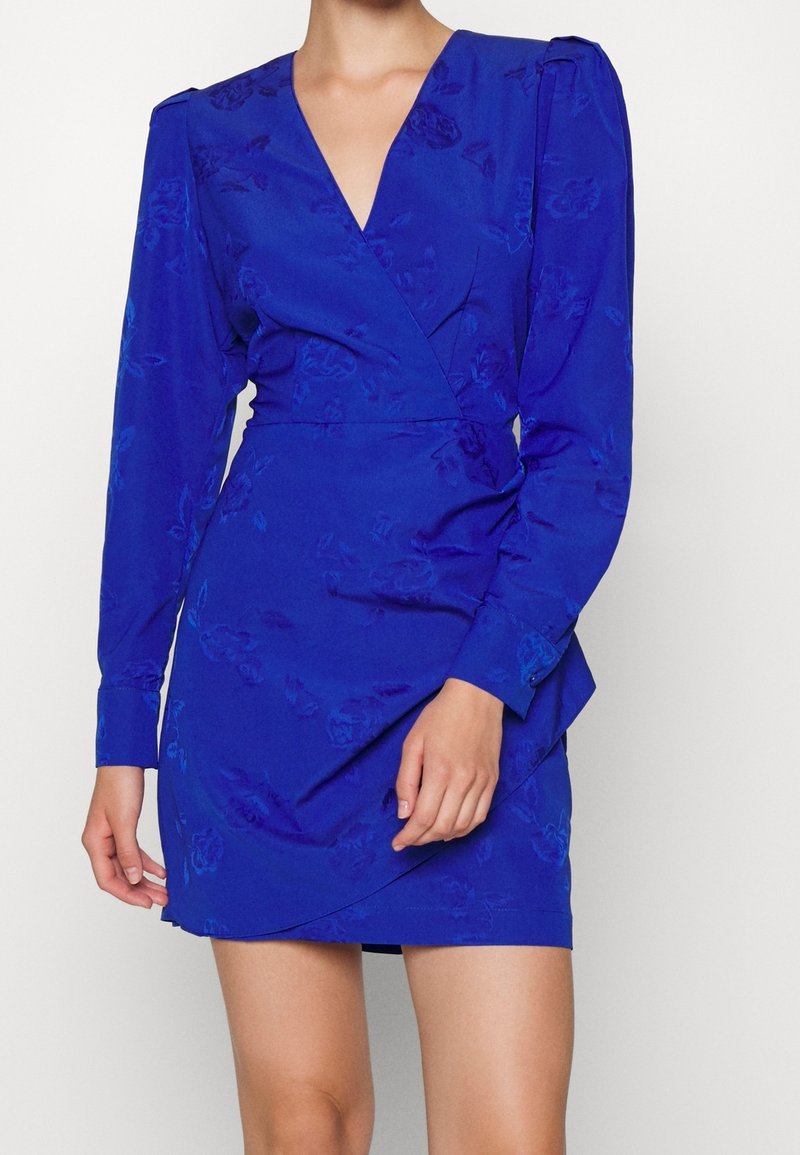Femme portant une robe portefeuille bleu roi à manches longues avec une texture florale subtile, un décolleté en V et une jupe courte cintrée.