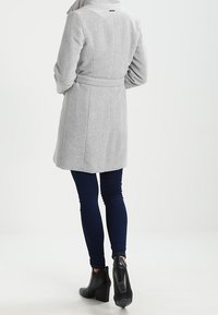 Abrigo gris con cintura ceñida, hombros estructurados y textura suave. Combinado con pantalones vaqueros ajustados de color azul oscuro y botines negros de tacón.