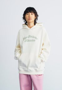 Hoodie crème avec "New Balance Athletics" en lettres vertes en relief. Comprend une poche kangourou et des poignets côtelés. Assorti avec un pantalon de survêtement rose.