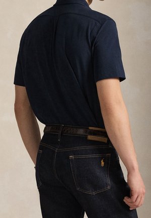 Mand iført mørkeblå kortærmet polo shirt, der er stoppet ned i mørke jeans med brunt bælte, stående mod en ensartet neutral baggrund.