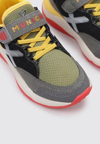 Zapatillas deportivas con un upper negro y detalles en verde olivo, cordones amarillos, suela gris y roja, y una correa que muestra "MUNICH."