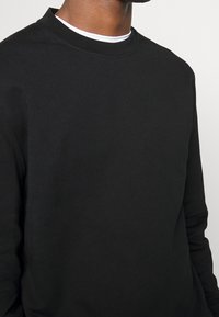 Felpa nera a collo rotondo, realizzata in tessuto texturizzato con maniche lunghe. Presenta un design semplice e un colletto spesso, sovrapposta a una camicia bianca sotto.