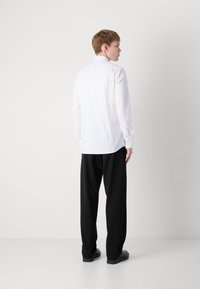 White long-sleeve shirt with a classic collar, button cuffs, and a straight hem, paired with black trousers, viewed from the back.