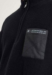 Schwarzer strukturierter Pullover mit einem hohen Kragen, ausgestattet mit einem schwarzen wattierten Patch auf der Brust, das weiße Texte und Logodetails zeigt.