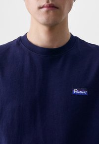 Tricou cu guler rotund de culoare bleumarin, fabricat din bumbac; prezintă un mic patch rectangular cu un logo pe pieptul stâng, având un design multicolor.