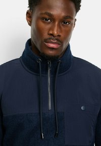 Marineblauer Pullover mit hohem Kragen, der eine Mischung aus glattem und strukturiertem Stoff aufweist, einen silbernen Reißverschluss und eine knöpfbare Brusttasche hat.