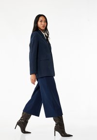 Marineblauw gestreepte blazer met twee knopen, gecombineerd met marineblauwe wijde broek. Model draagt kniehoge bruine puntige laarzen en staat op een effen achtergrond.