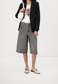 Femme portant un blazer noir, un t-shirt blanc avec un motif de nœud rouge, un short en denim gris large atteignant les genoux, des ballerines beiges pointues avec des lanières, et portant un sac blanc.
