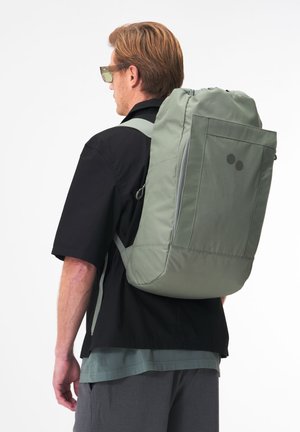Zaino verde imbottito con tasca frontale con cerniera, spallacci regolabili e design minimalista. Indossato sulla schiena di una persona in abbigliamento casual.