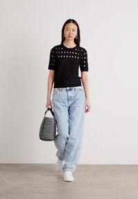 Black knit top with cut-out patterns, paired with light blue loose jeans and white sneakers. The model carries a textured gray bucket bag.