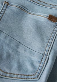 Lyseblå denimstoff med en synlig baklomme, detaljerte sømmer og et brunt lærmerke for merkevarebygging. Jevn tekstur.