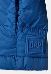 Modrá prošívaná bunda s lesklou látkou a obdélníkovou gumovou nášivkou s logem GAP na levé straně.
