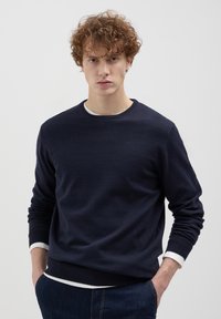 Marineblauer Pullover aus weichem Strickstoff mit Rundhalsausschnitt, gerippten Bündchen und Saum, über einem weißen Hemd getragen.