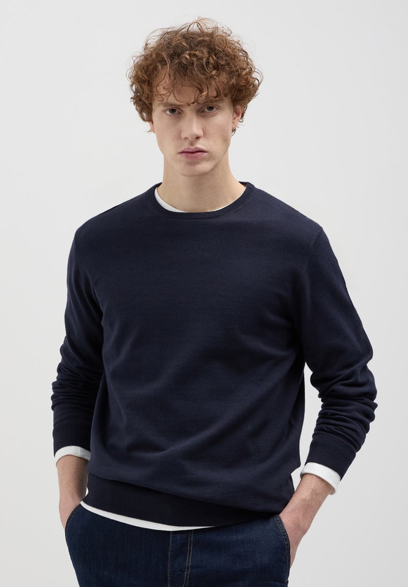 Marineblauer Pullover aus weichem Strickstoff mit Rundhalsausschnitt, gerippten Bündchen und Saum, über einem weißen Hemd getragen.