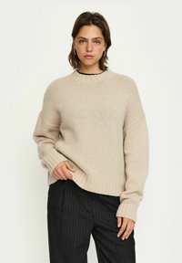 Beige gestrickter Pullover mit Rundhalsausschnitt, tiefen Schultern und gerippten Bündchen. Kombiniert mit schwarzen Nadelstreifen-Hosen.