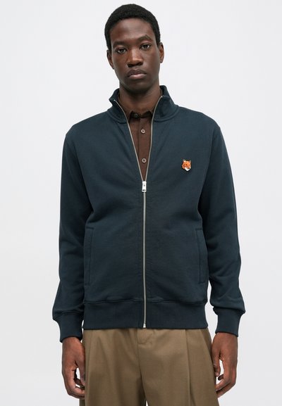 Uomo che indossa una giacca con zip blu navy con un logo a forma di testa di volpe sul petto, una camicia marrone sotto e pantaloni beige su uno sfondo bianco.