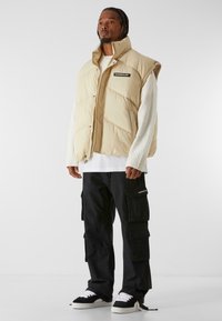 Beige padded vest with a high collar and logo patch, worn over a white sweater and black cargo pants with multiple pockets.
