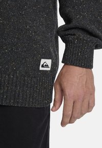 Schwarz gesprenkelter Strickpullover mit gerippten Ärmeln. Verfügt über ein kleines weißes Stoffetikett mit einem Berglogo nahe dem Saum.
