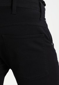 Svarta jeans har en figurnära design med en synlig bakficka och platta sömmar. Tyget har en slät, matt yta.