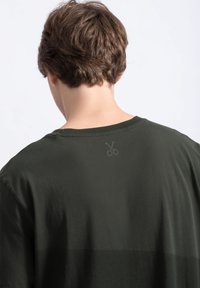 Donkergroene katoenen t-shirt met een ontspannen pasvorm. Heeft een subtiele schaarafbeelding op de bovenrug gedrukt. Vlotte textuur.