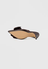 Zapato de tacón alto marrón con puntera puntiaguda y detalle de lazo. Presenta un acabado de charol elegante y una suela de goma nude con agarre texturizado.