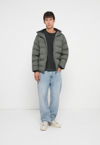 Φούτερ ζακέτα σε σκούρο πράσινο, με κλείσιμο με φερμουάρ, κουκούλα και ραμμένη σχεδίαση. Συνδυάζεται με ελαφρώς εφαρμοστά ανοιχτούχρωμα μπλου τζιν και λευκά παπούτσια.