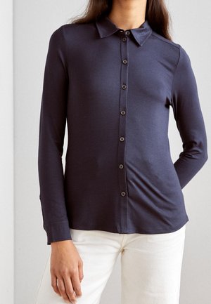 Vrouw draagt een marineblauw overhemd met lange mouwen en witte broek, staat met één hand ontspannen langs haar zijde.