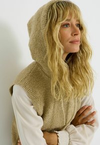 Gilet à capuche beige en tricot avec des manches courtes, tissu texturé et finition douce. Porté sur une blouse blanche avec des poignets à volants.