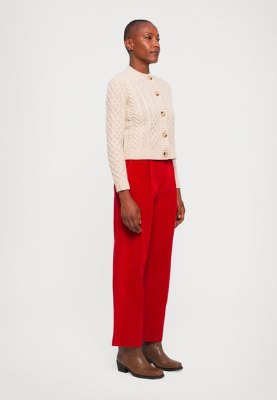 Beige cable-knit cardigan with gold buttons, paired with high-waisted red corduroy pants and brown ankle boots, displayed against a neutral background.
