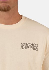 Beige puuvillane t-särk, millel on must "WRONG FRIENDS" logo vasakul rinnal. Kangas on sileda tekstuuriga ja klassikalise ümarkaelusega disain.