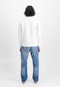 En person med krøllete, skulderlangt hår står med ryggen til, iført en enkel hvit hettegenser, blå jeans og svart-hvite sneakers.