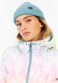 Mehrfarbige Daunenjacke mit einem Farbverlaufsmuster (lila zu pink), ausgestattet mit einer Kapuze und einem türkisfarbenen Reißverschluss; kombiniert mit einer hellblauen gerippten Mütze.